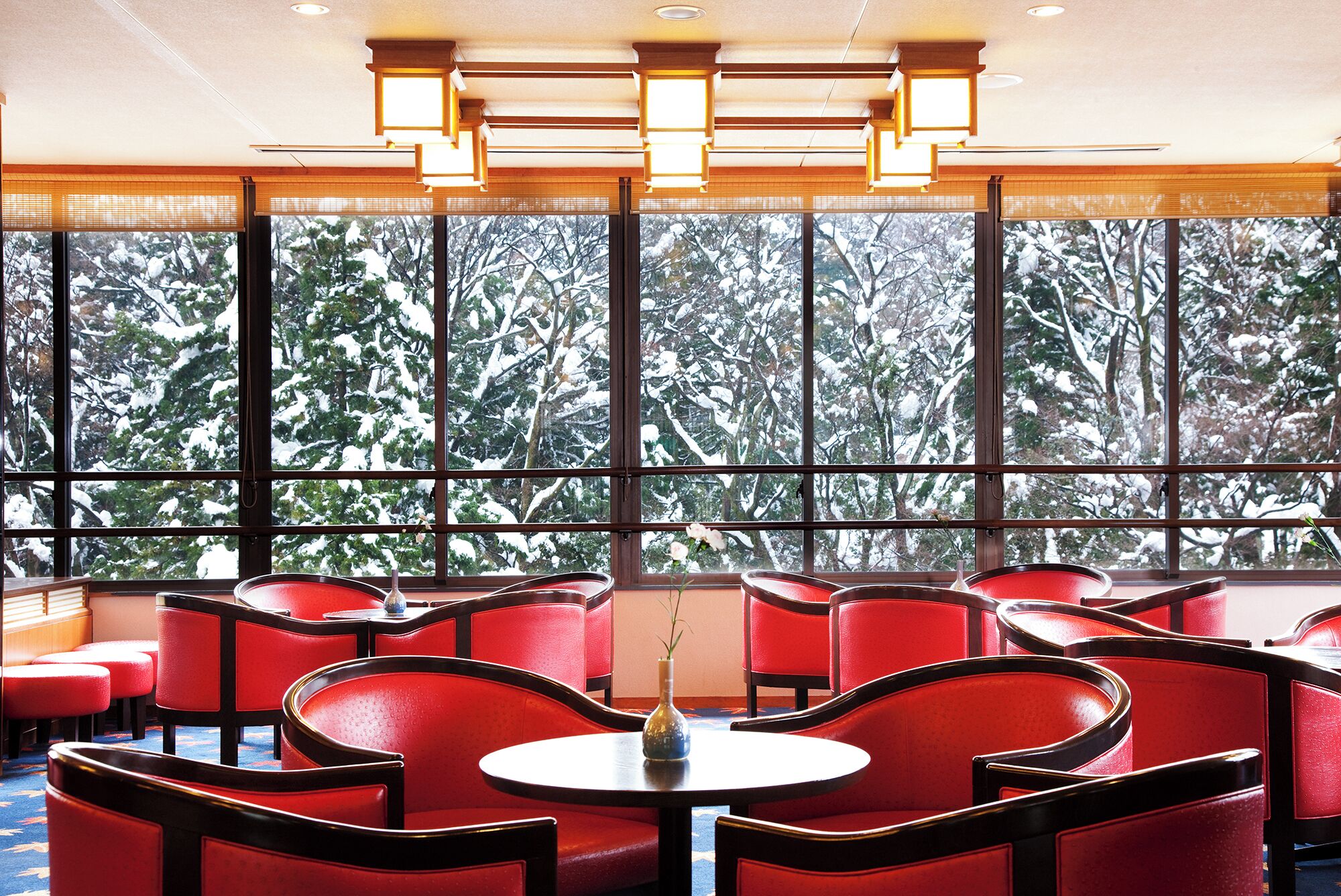Habitación de hotel con ventanas amplias, sillones tapizados en rojo y una mesa redonda.