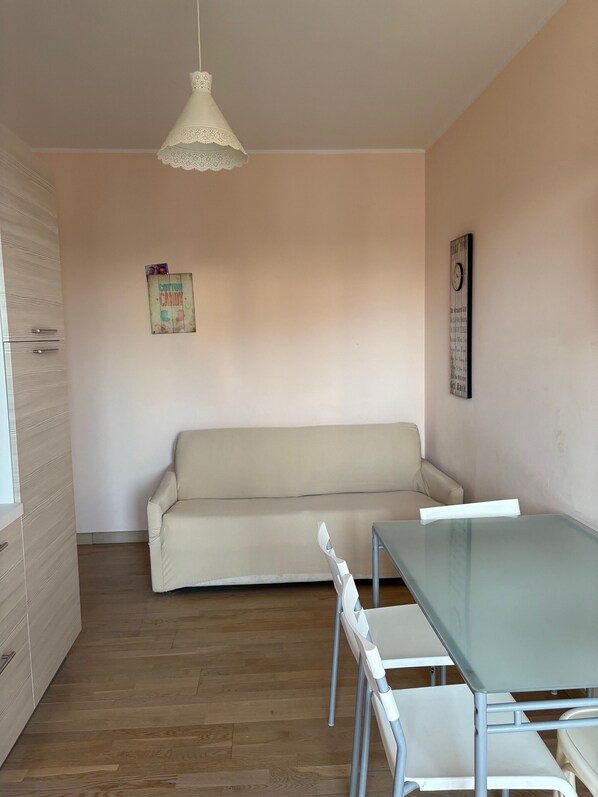 A room with a glass table, white chairs, a beige sofa, a white pendant light, and a wall clock. - GRADO - BEAUTIFUL ONE BEDROOM APARTMENT WITH LARGE BALCONY (Grado)