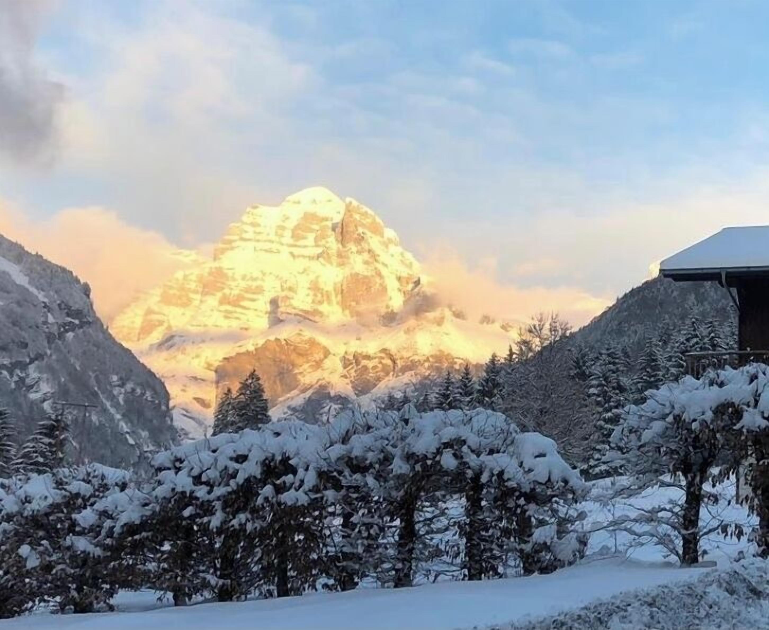 Une montagne enneigée, avec un sommet bien visible, entourée d’arbres et d’un bâtiment.