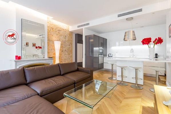 A modern living room with a brown leather sofa, a glass coffee table, and a kitchen area featuring a white countertop, stainless steel appliances, and red flowers.