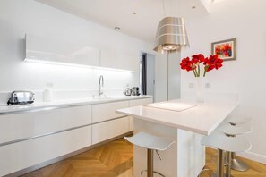 A modern kitchen with white cabinetry, a central island with bar stools, a pendant light, and a vase with red flowers on the wall.