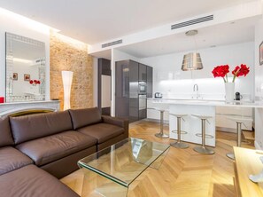 Un salon moderne avec un canapé en cuir marron, une table basse en verre et un coin cuisine doté d’un plan de travail blanc, d’appareils électroménagers en acier inoxydable et de fleurs rouges.