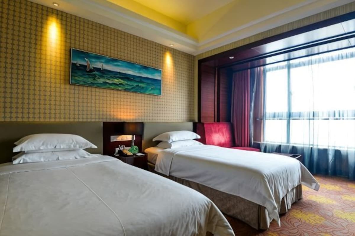 Quarto de hotel com duas camas, um quadro na parede, uma janela grande com cortinas e um tapete estampado.