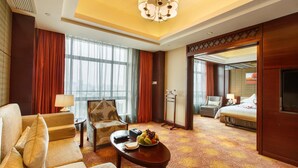 A hotel room with a sofa, armchair, and a dining table with fruits. There is a bed with a TV and a large window with curtains. - Leeden Jingxi Hotel (Guangzhou)