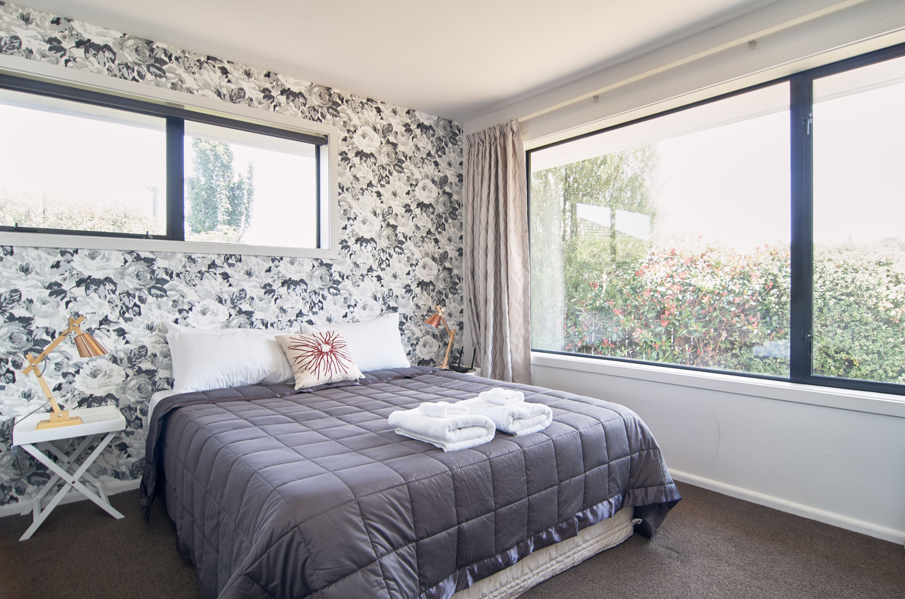 A bedroom with a large bed, floral wallpaper, and a view of greenery through a window.
