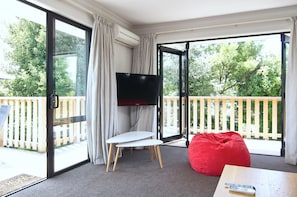 A room with a sliding glass door leading to a balcony, a flat-screen TV mounted on the wall, a small white table, a red bean bag, and a remote control on a wooden table. - Brookside Villa (Christchurch)