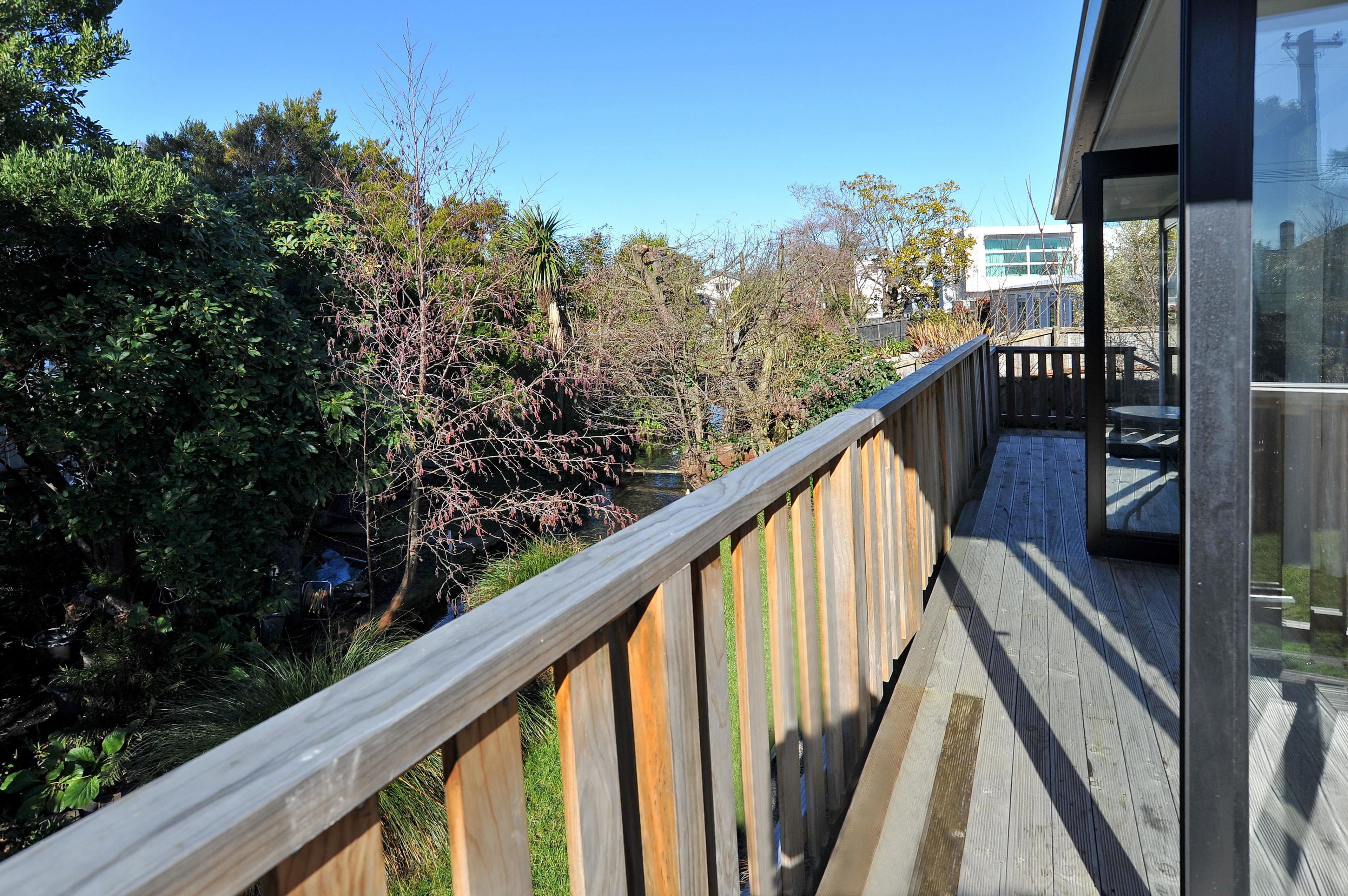 A wooden deck with a glass railing overlooking a garden and a building in the background.