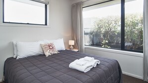 A neatly made bed with a dark quilt, white pillows, and a decorative pillow. There is a small bedside table with a lamp and a window with curtains. - Brookside Villa (Christchurch)