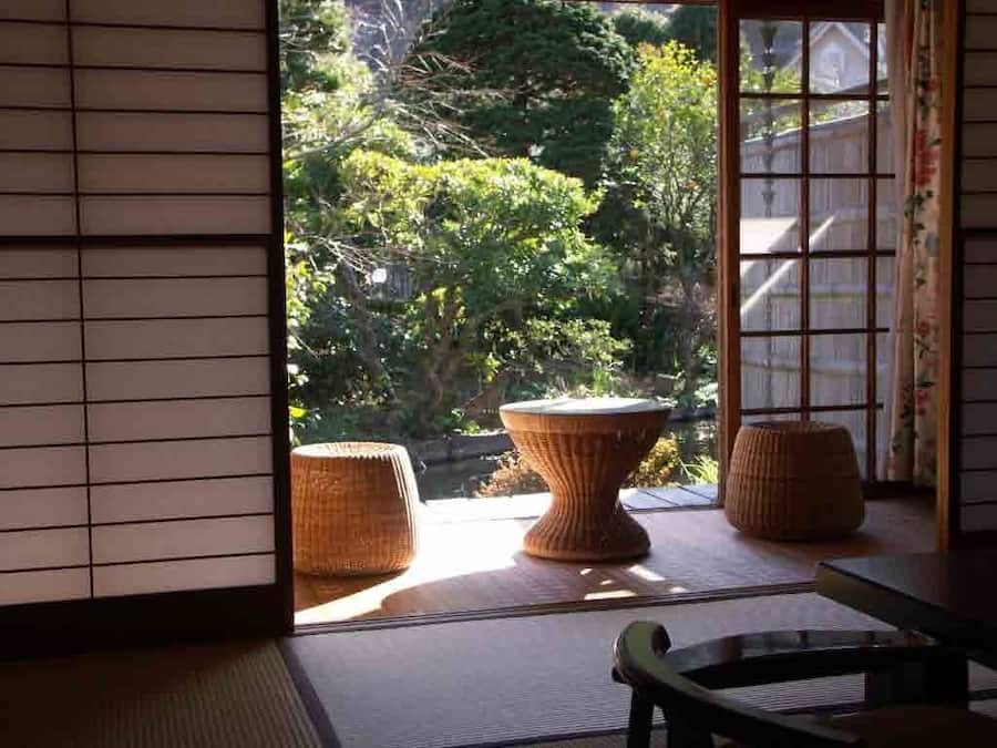 A traditional Japanese room with tatami flooring, a low table, and a view of a garden with a pond and trees.