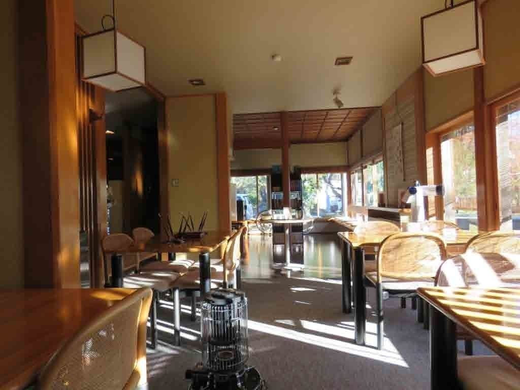 A spacious restaurant with high ceilings and natural light from large windows. Tables are arranged in a casual layout, and there are chairs with woven seats.