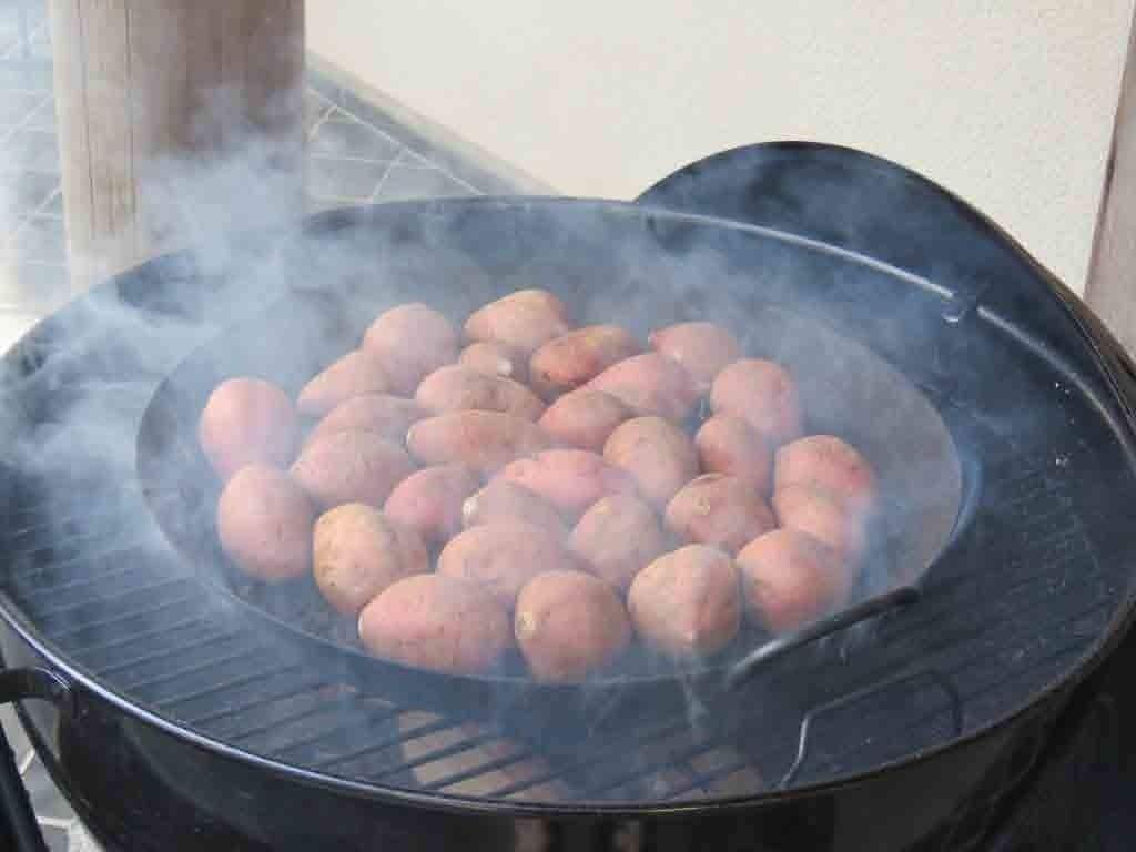 Un barbecue extérieur de forme ronde sur lequel sont en train de cuire plusieurs pommes de terre.