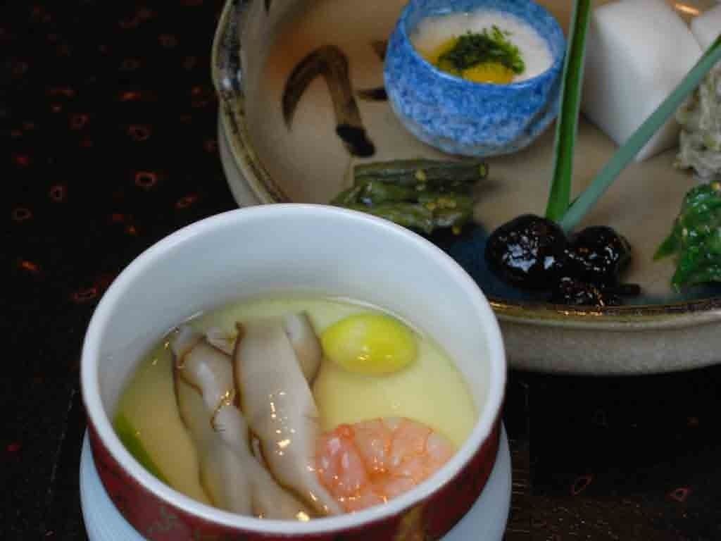A bowl of soup with various ingredients, a small bowl of sauce, and some vegetables on a table.