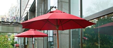An outdoor seating area with tables and chairs, red umbrellas, and a view of a building with large windows.