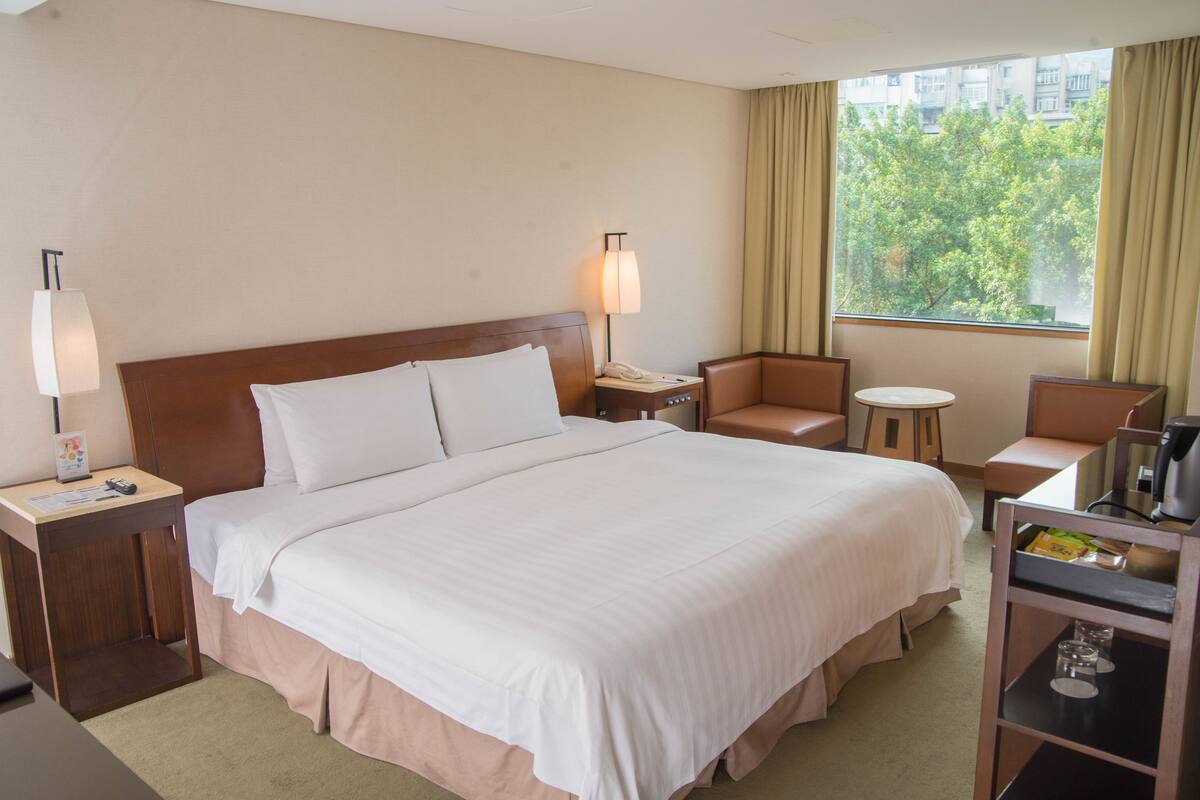 A hotel room with a large bed, a bedside table with a lamp, a chair, a small table, and a window with curtains.