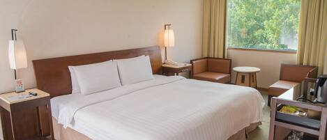 A hotel room with a large bed, a bedside table with a lamp, a chair, a small table, and a window with curtains.