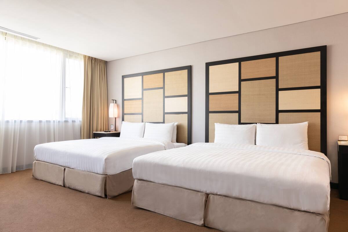 A hotel room with two beds, a large window with curtains, and a wall-mounted light fixture.