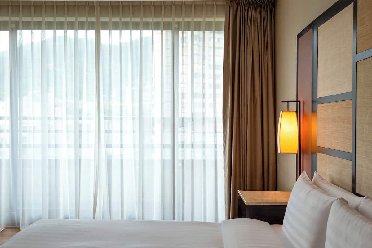 A hotel room with a large bed, a nightstand with a lamp, sheer curtains, and a view of buildings through the window.