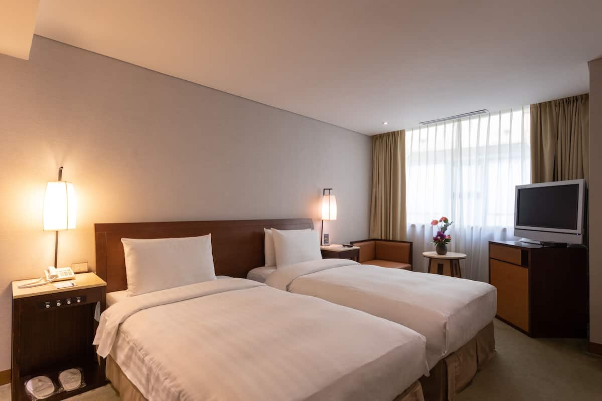A hotel room with two beds, a TV, a desk with a lamp, and a small table with a vase.