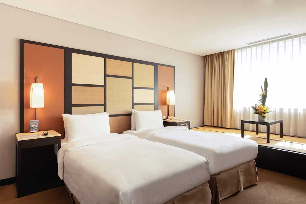 A hotel room with two beds, a large window with curtains, a bedside table with a lamp, and a small table with a vase of flowers.