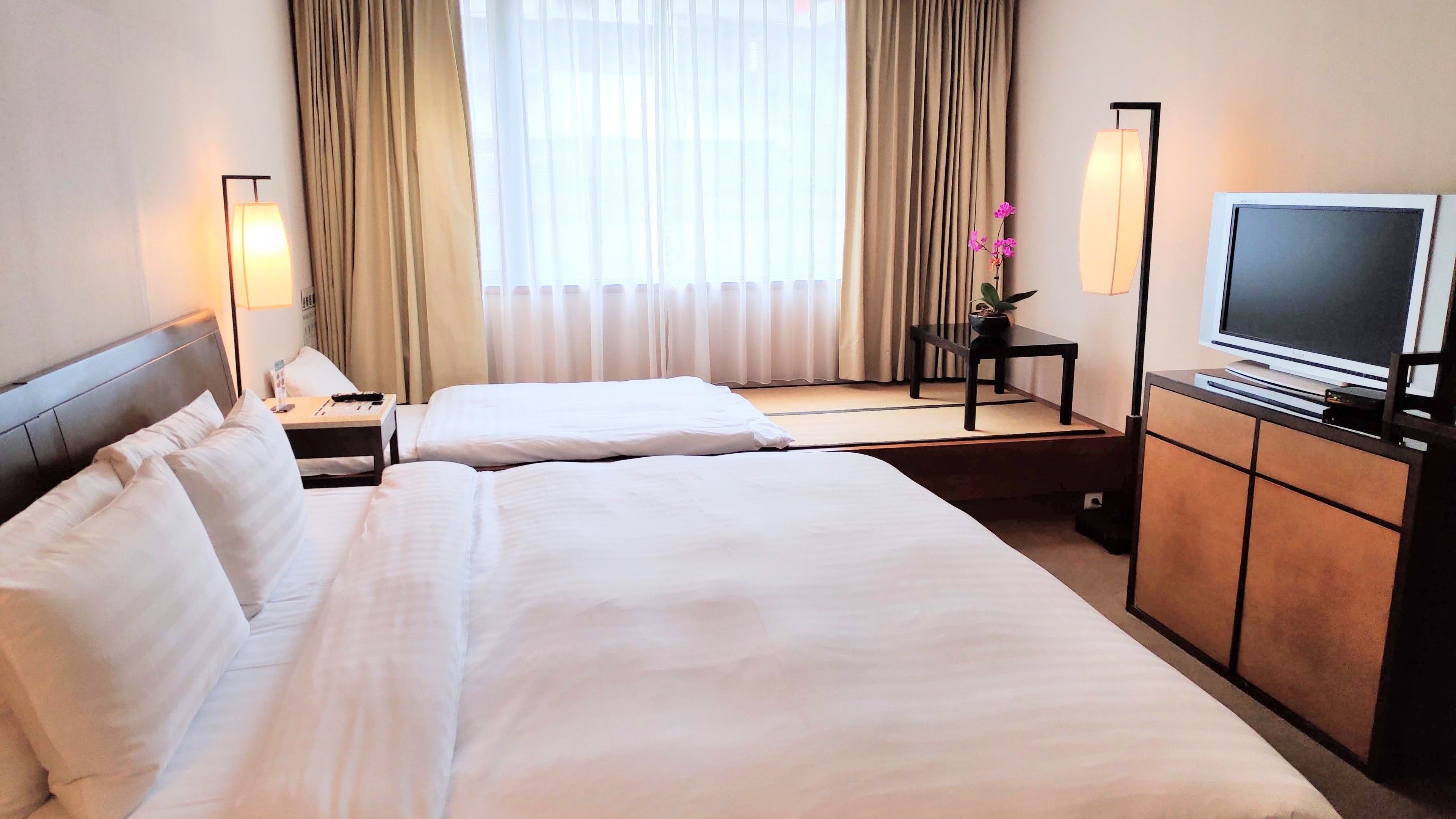 A hotel room with two beds, a TV, a bedside table with a lamp, and a window with curtains.