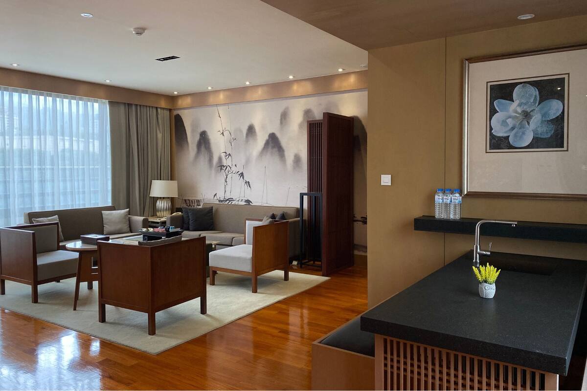 A modern hotel room with a sofa set, a coffee table, a desk, a small kitchenette, and a large window with curtains.