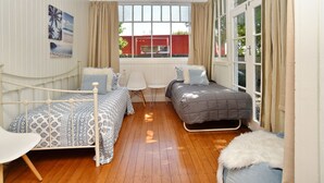A bedroom with two beds, a white chair, a small table, and a large window with curtains. - Racecourse Villa (Christchurch)