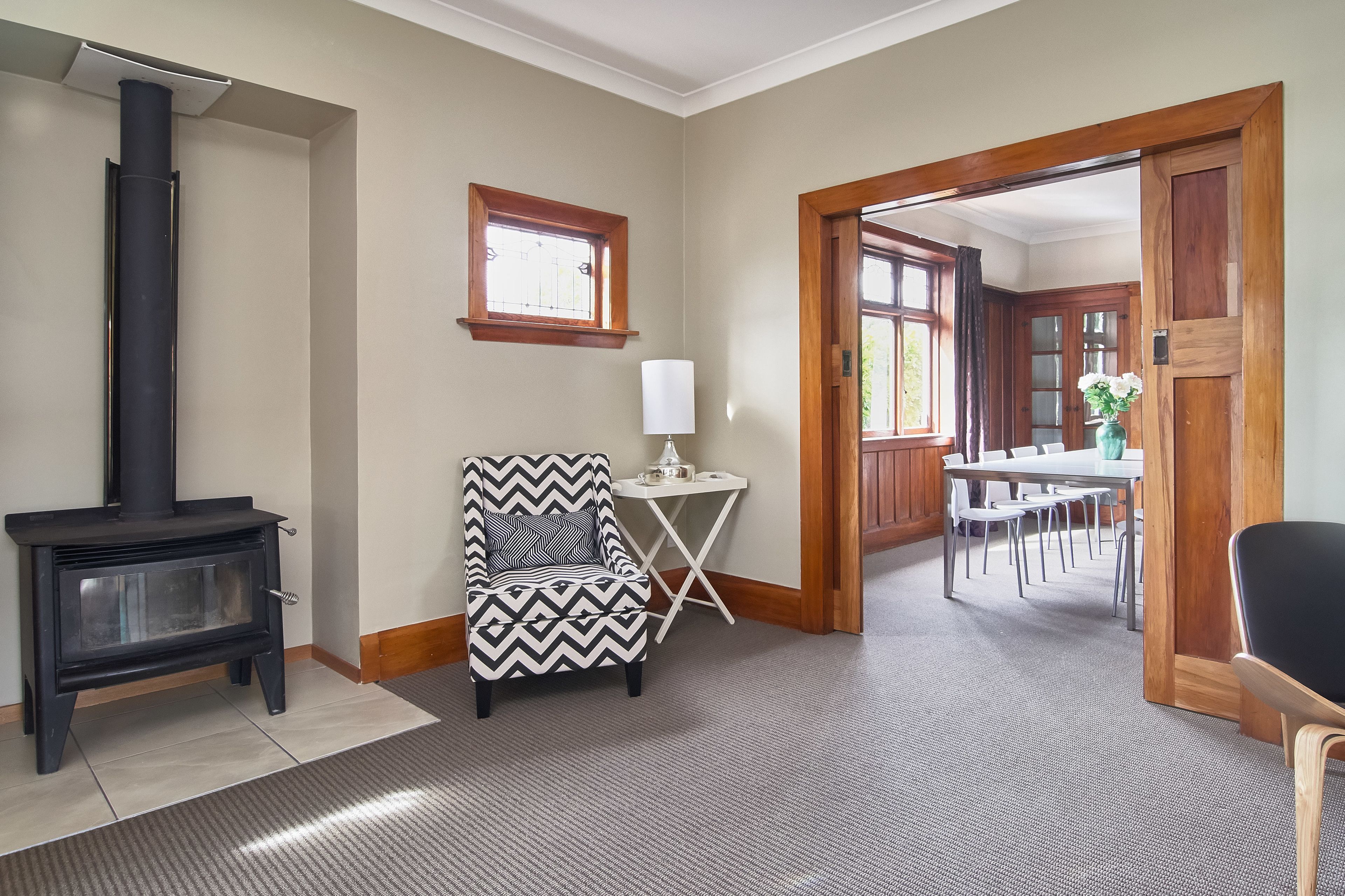 A room with a black wood-burning stove, a patterned armchair, a small white table with a lamp, and a dining area visible through an open doorway.