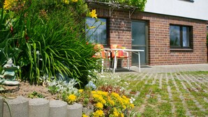 A modern house with a brick exterior, a small garden with various plants and flowers, and a patio area with a table and chairs. - "Bergblick" guest apartment near Löbauer Berg (Upper Lusatia) (Löbau)