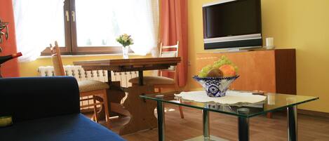 Ein Wohnzimmer mit Glastisch, einer Couch, einem Esstisch mit Stühlen, einem Fernseher und einem Fenster mit Vorhängen.