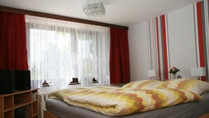 A bedroom with a bed, a radiator, a TV on a shelf, and a window with curtains. - "Bergblick" guest apartment near Löbauer Berg (Upper Lusatia) (Löbau)