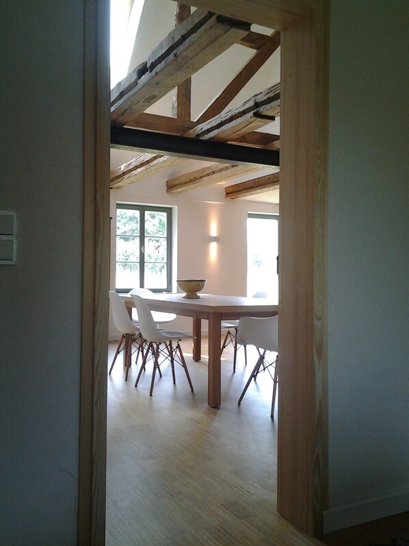 An open-plan dining area with a wooden table and white chairs, a large window with a view of greenery, and exposed wooden beams on the ceiling.