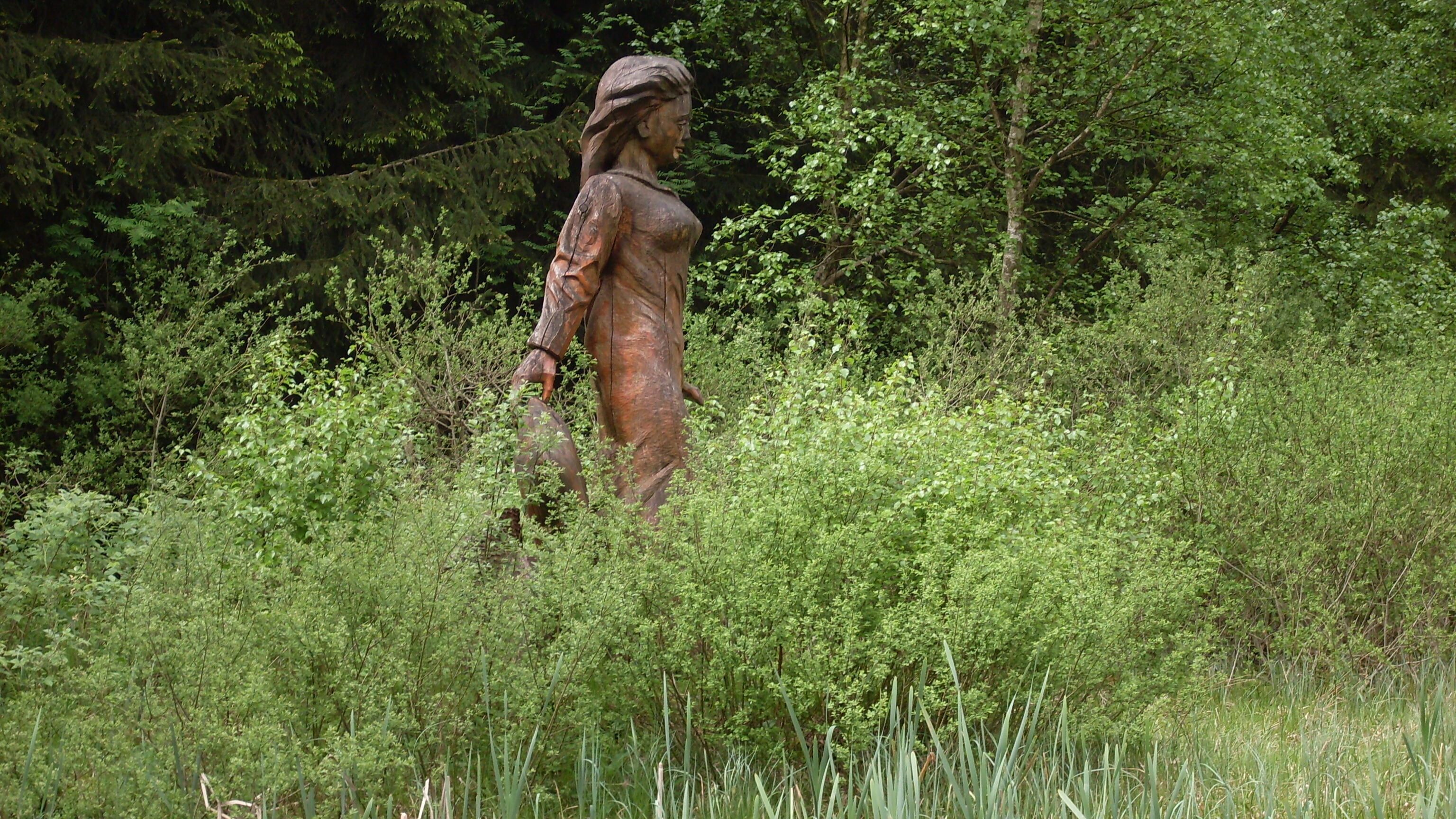 A statue of a woman in a natural setting with dense foliage and trees in the background.