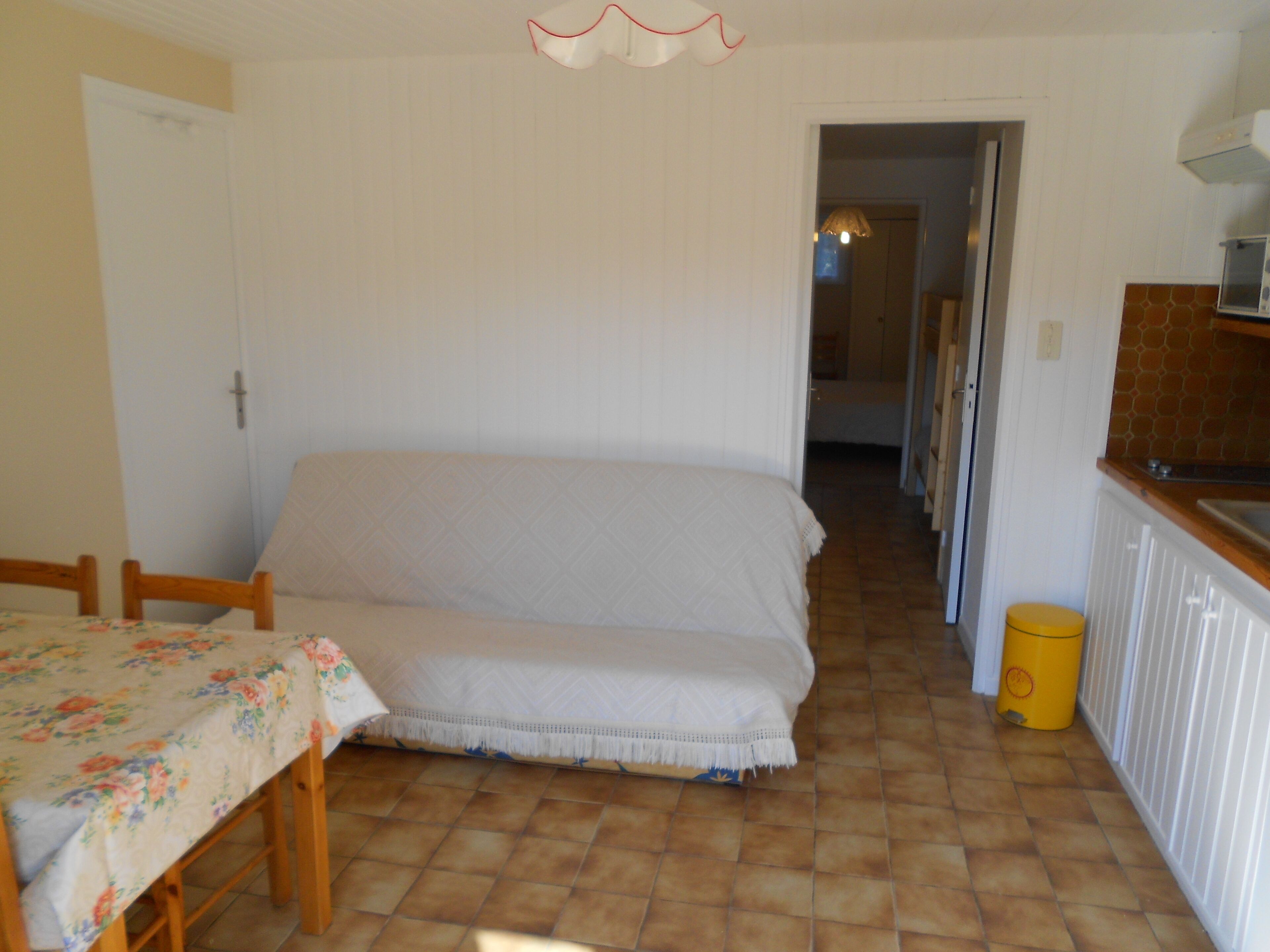 Un dormitorio con una cama, una mesa con mantel floral, un bote de basura amarillo y una zona de cocina con microondas.