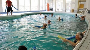 An indoor swimming pool with clear water, a lifeguard, and people swimming and using floatation devices.