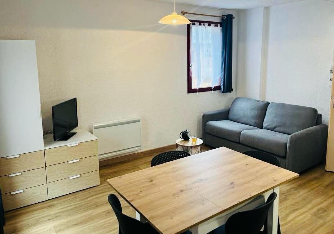 Une petite pièce bien éclairée, avec une table en bois, un canapé gris, un téléviseur posé sur un meuble et une fenêtre avec des rideaux.