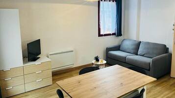 Une petite pièce bien éclairée, avec une table en bois, un canapé gris, un téléviseur posé sur un meuble et une fenêtre avec des rideaux.