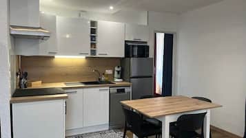 Een compacte keuken met witte kasten, een houten aanrecht, een spoelbak, een kookplaat en een koelkast. Er staat een eettafel met stoelen.