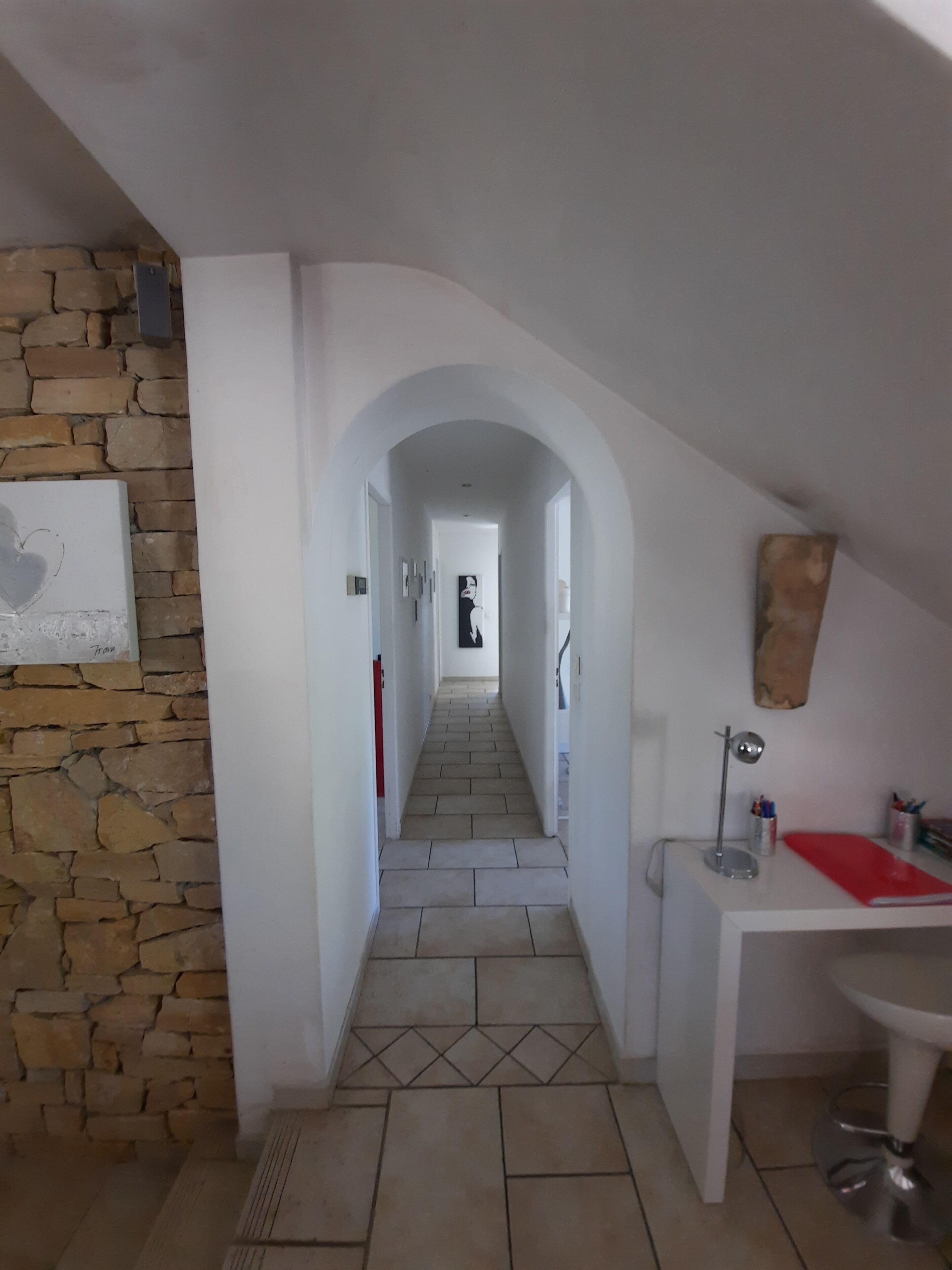 A hallway with a stone wall on one side, a tiled floor, and a desk with a red notebook and pens on the right.