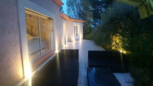 Ein überdachter Außenbereich mit Terrasse, einer Tür zu einem überdachten Gang und Blick auf den umliegenden Garten.