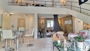 Ein geräumiger Wohnbereich mit hoher Decke, einer Steinwand und einem Kamin. Es gibt einen Essbereich mit einem Glastisch und Stühlen.