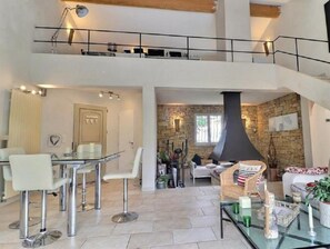 Un amplio salón con techo alto, pared de piedra y chimenea. Hay un comedor con mesa y sillas de vidrio.