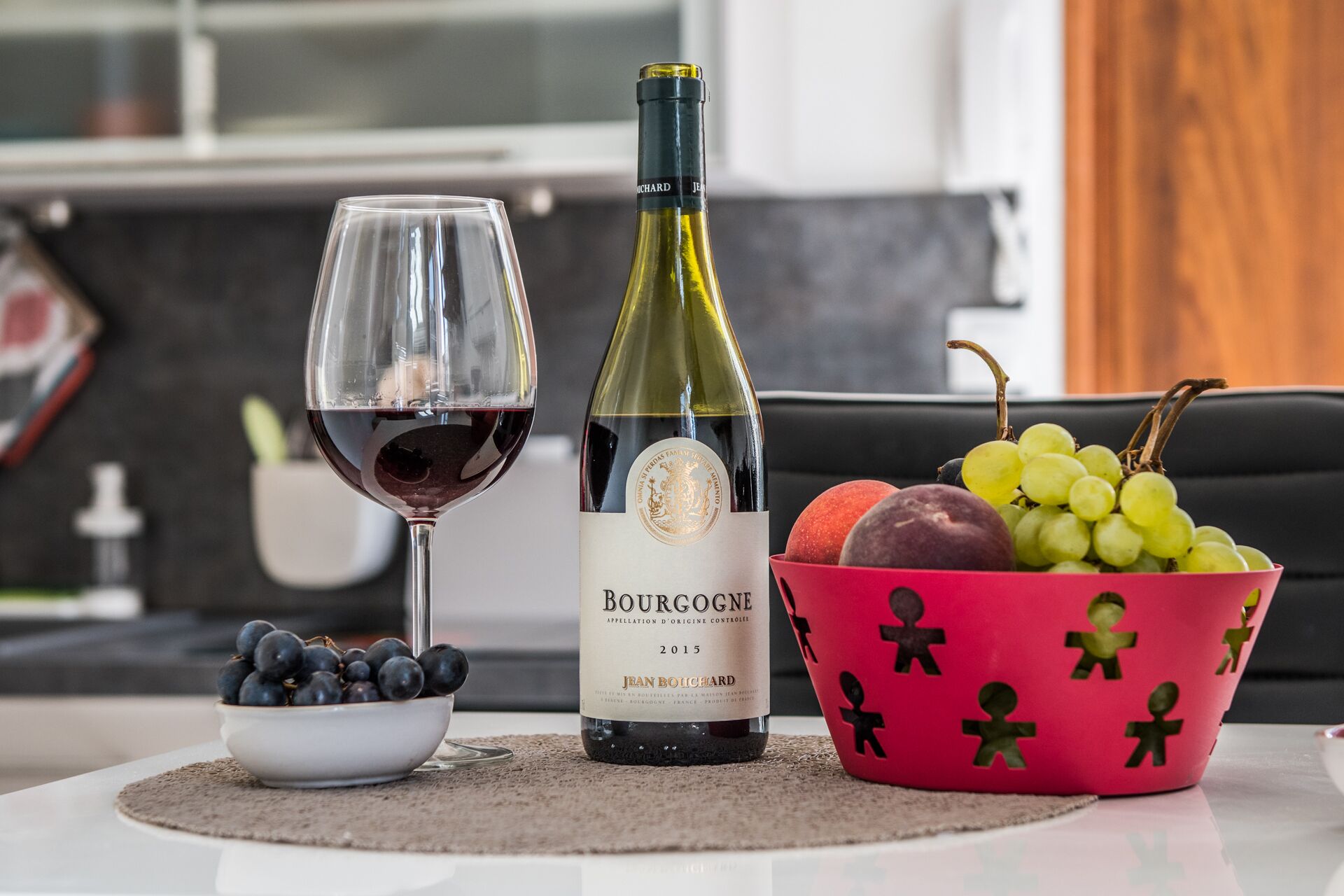 Une bouteille de vin de Bourgogne, un verre de vin rouge et un bol de raisins sur une table.
