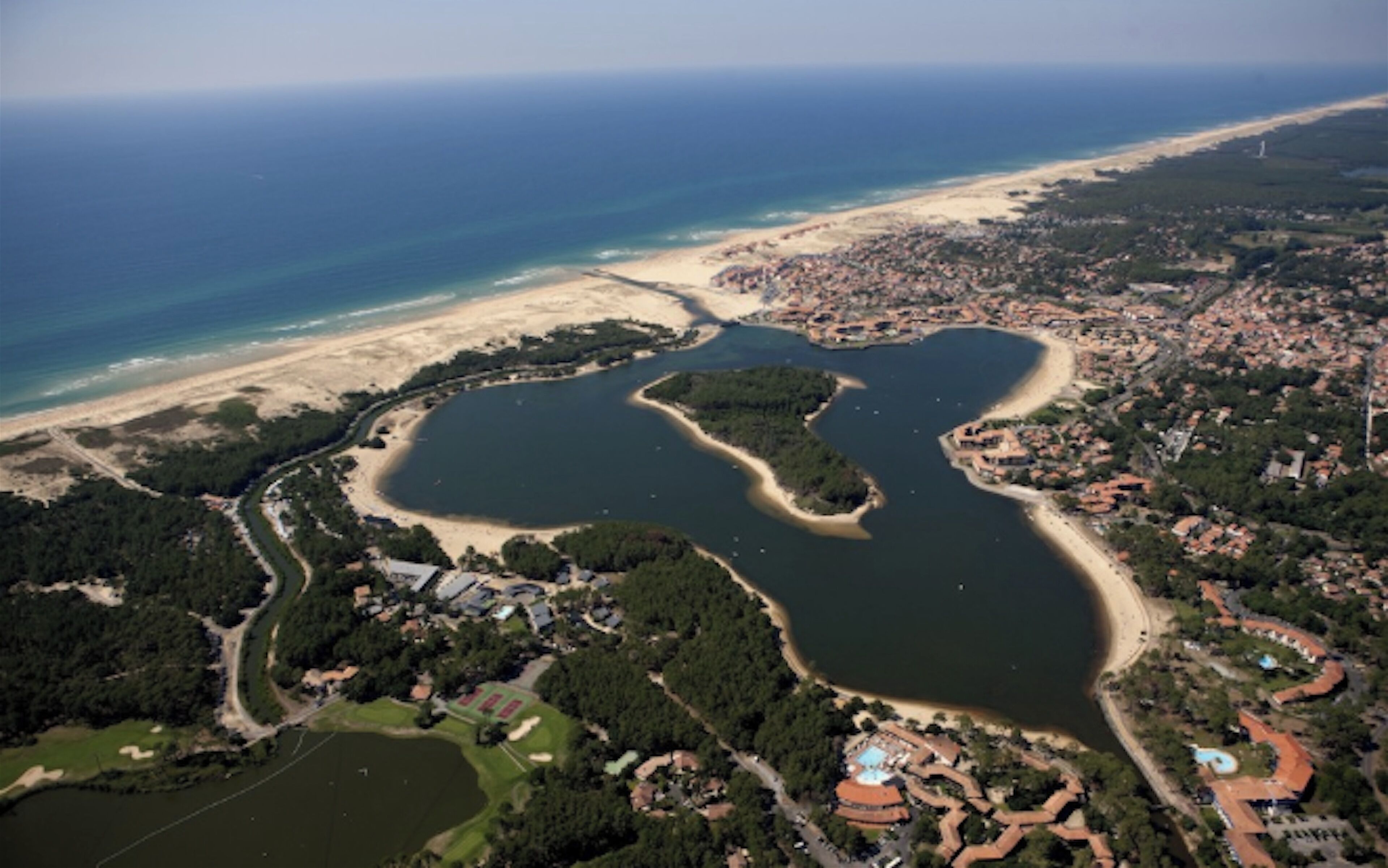 An aerial view of a coastal area with a beach, a lake, and a residential area with houses and greenery.