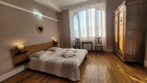 Un dormitorio con cama de madera, lámparas de noche, una mesita, dos sillas, un armario y una ventana con cortinas traslúcidas.