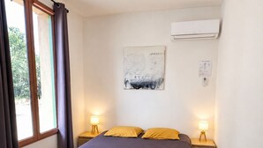 A hotel room with a bed, two bedside lamps, an air conditioner, a painting on the wall, and a window with curtains. - Quiet Salagou cottage with lovely view over the valley (Octon)