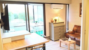 A compact living space with a sliding glass door leading to a balcony, a small table with a laptop, a leather sofa, and a cabinet with drawers. - Quiet air-conditioned apartment 10 minutes' walk from the beaches and shops (Bormes-Les-Mimosas)