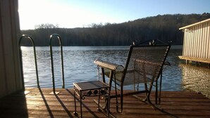 A wooden deck with a metal chair and a small table, overlooking a calm lake with a forested shoreline.