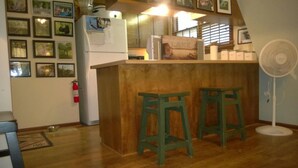 A kitchen with a wooden island, two green stools, a white refrigerator, and a white fan.