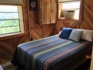 A wooden cabin bedroom with a bed, pillows, a window, and an air conditioning unit.