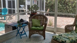 A sunroom with a view of a backyard, a wicker chair with a green cushion, a blue table, and a potted plant. - Cosy Home with Heated Pool (Edgewater)